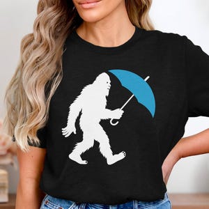 Può includere: T-shirt nera con grafica di Bigfoot bianco che tiene un ombrello blu. Il design raffigura la leggendaria creatura che cammina con l'ombrello aperto. Un design divertente per i fan dei criptidi.