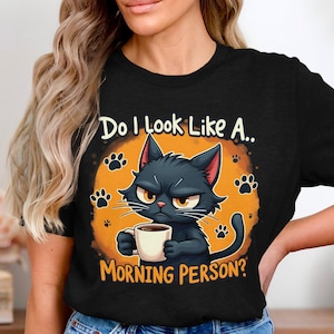 Puede incluir: Camiseta negra con un gato negro de dibujos animados sosteniendo una taza de café. El texto en la camiseta dice "Do I look like a... Morning Person?"