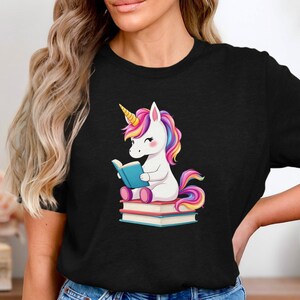 Könnte beinhalten: Schwarzes T-Shirt mit einem Cartoon-Einhorn-Design. Das Einhorn mit gelbem Horn, Regenbogenmähne und -schwanz liest ein blaues Buch, während es auf einem Stapel Bücher sitzt. Das Einhorn hat rosa Wangen und lächelt.