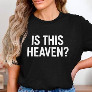 Puede incluir: Camiseta negra con el texto blanco "IS THIS HEAVEN?". La camiseta es de cuello redondo. La imagen muestra la camiseta en una persona.