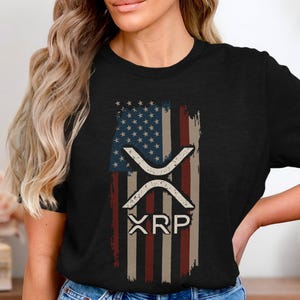 May include: Black t-shirt featuring a distressed American flag design with the XRP cryptocurrency symbol and the word "XRP" in white. The flag design incorporates blue, red, and white colors, creating a patriotic theme.