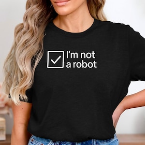 Op de afbeelding: Zwart T-shirt met de witte tekst "I'm not a robot". De tekst staat naast een witte vierkant met een vinkje. Het T-shirt heeft een ronde hals en korte mouwen.