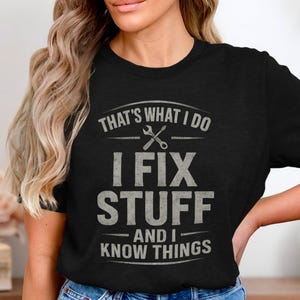 May include: Black t-shirt with the text "THAT'S WHAT I DO I FIX STUFF AND I KNOW THINGS" in a distressed, gray font. The design includes crossed wrenches above the text. The shirt is a crew neck style.