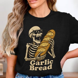 Könnte beinhalten: Schwarzes T-Shirt mit einem Skelett, das eine Sonnenbrille trägt und ein Knoblauchbrot hält. Die Worte "Garlic Bread" sind unter dem Skelett in Beige gedruckt. Das Design ist eine humorvolle Interpretation von Essen.