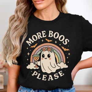Könnte beinhalten: Schwarzes T-Shirt mit einem Cartoon-Geist, der einen Cocktail hält, einem Regenbogen und dem Text "MORE BOOS PLEASE". Das Design umfasst Fledermäuse und Sterne, passend für Halloween.