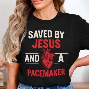 Könnte beinhalten: Schwarzes T-Shirt mit dem Text "SAVED BY JESUS AND A PACEMAKER" in Weiß und Rot. Eine rote anatomische Herzgrafik ist zwischen den Sätzen zentriert. Das Shirt hat einen Rundhalsausschnitt.