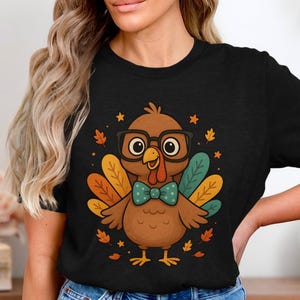 May include: Black t-shirt featuring a cartoon turkey wearing glasses and a bow tie. The turkey has colorful feathers and is surrounded by autumn leaves and stars. A fun, festive design.