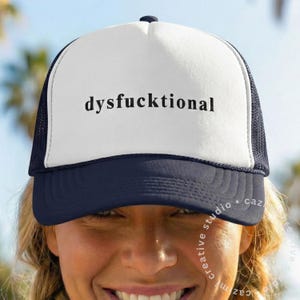 May include: A white trucker hat with a navy blue brim and mesh back. The hat features the word "dysf*cktional" in black, centered on the front panel. The hat is worn by a person with blonde hair.