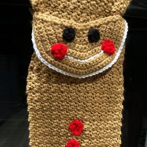 May include: Crocheted gingerbread man ornament with red and white details. The ornament is brown and has a white trim around the edges.
