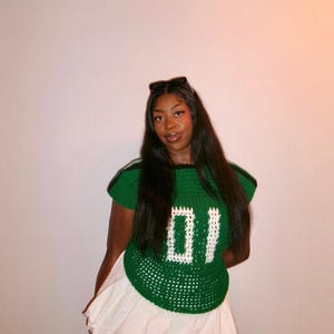 May include: A green crochet top with the number "01" in white, paired with a white skirt. The top has green and white stripes on the shoulders. The outfit is a unique, handmade fashion statement.