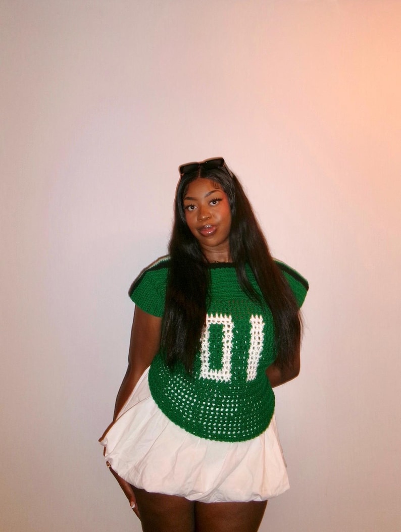 May include: A green crochet top with the number "01" in white, paired with a white skirt. The top has green and white stripes on the shoulders. The outfit is a unique, handmade fashion statement.