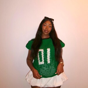 May include: A green crochet top with the number "01" in white, paired with a white skirt. The top has a round neckline and short sleeves. Sunglasses are perched on top of the head.