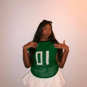 May include: A green crochet top with the number "01" in white, paired with a white bubble skirt. The top has a round neckline and short sleeves. The outfit is a unique, handmade fashion statement.