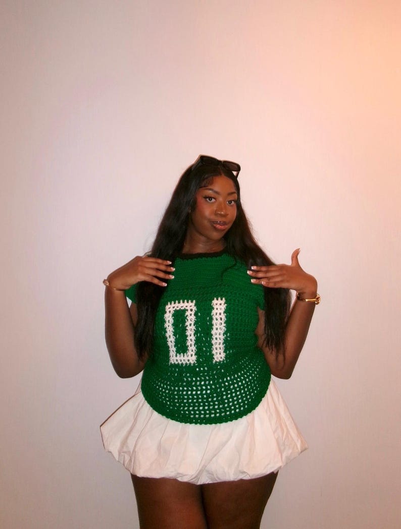 May include: A green crochet top with the number "01" in white, paired with a white bubble skirt. The top has a round neckline and short sleeves. The outfit is a unique, handmade fashion statement.