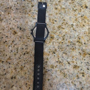 May include: Black wristwatch with a black nylon strap. The watch has a black face and a silver buckle. The strap has multiple adjustment holes. The watch is lying on a speckled surface.