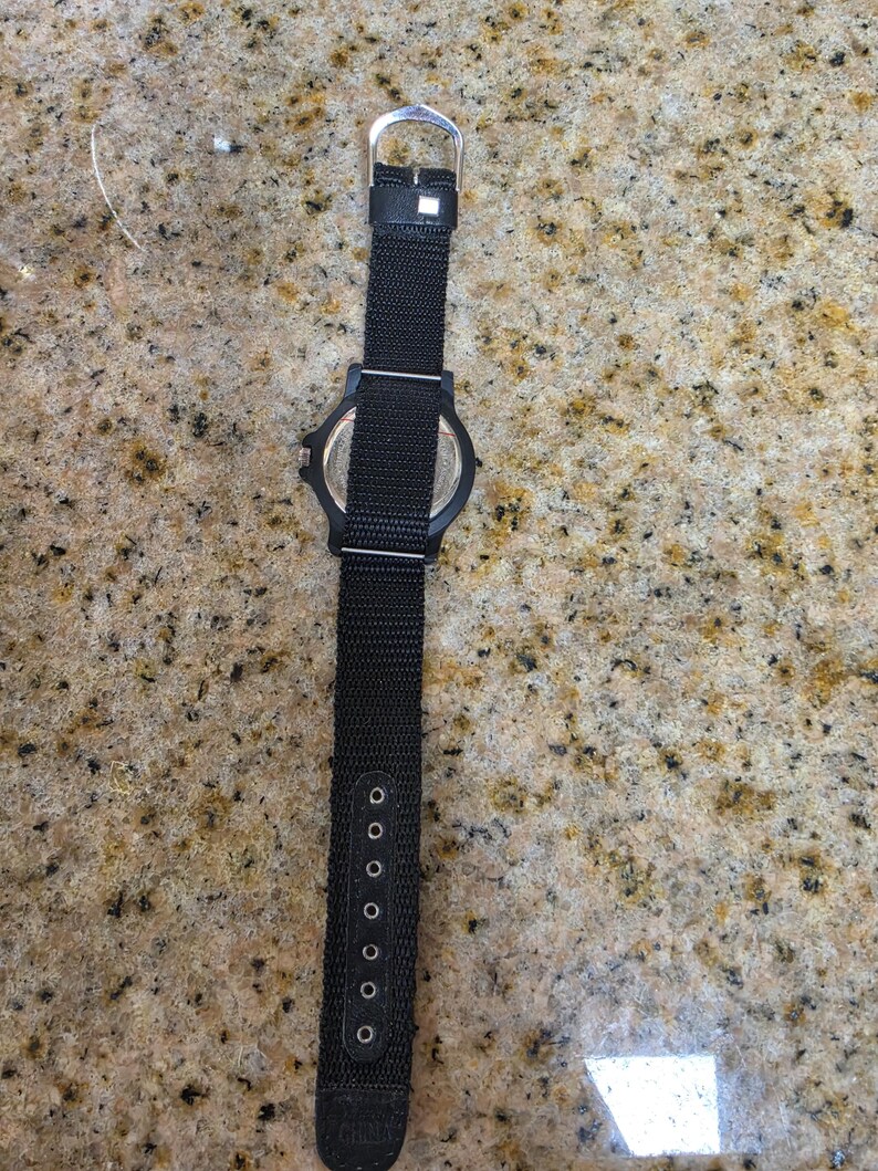 May include: Black wristwatch with a black nylon strap. The watch has a black face and a silver buckle. The strap has multiple adjustment holes. The watch is lying on a speckled surface.