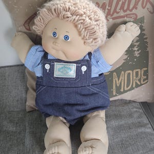 May include: A vintage Cabbage Patch Kids doll with light skin, blue eyes, and light brown yarn hair. The doll wears a blue gingham shirt under denim overalls with white buttons. A Cabbage Patch Kids label is sewn on the front.