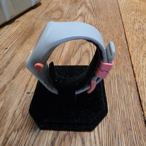 May include: A gray wristwatch with a pink clasp and a red button. The watch is displayed on a black velvet stand. The watch has a modern, minimalist design.