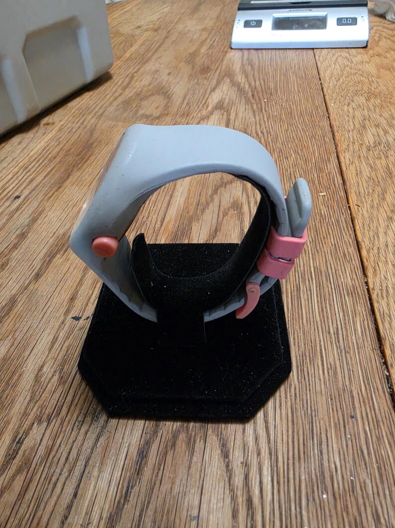 May include: A gray wristwatch with a pink clasp and a red button. The watch is displayed on a black velvet stand. The watch has a modern, minimalist design.