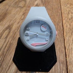 May include: A gray Puma watch with a round face and a gray silicone band. The watch face features white numerals, white hands, and a pink Puma logo. The watch is displayed on a black velvet stand.