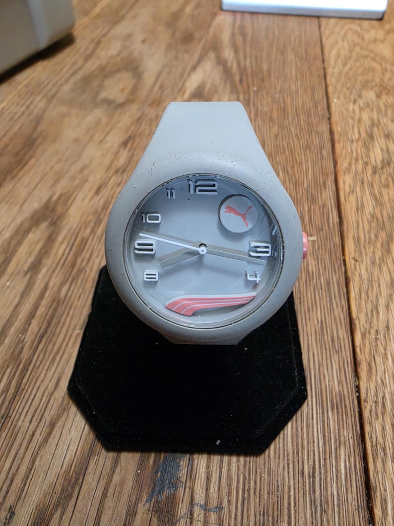 May include: A gray Puma watch with a round face and a gray silicone band. The watch face features white numerals, white hands, and a pink Puma logo. The watch is displayed on a black velvet stand.