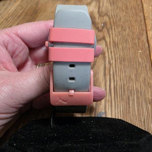 May include: A gray watch band with a pink clasp and straps. The watch band is held over a black velvet display pillow. The watch band is made of a flexible material.