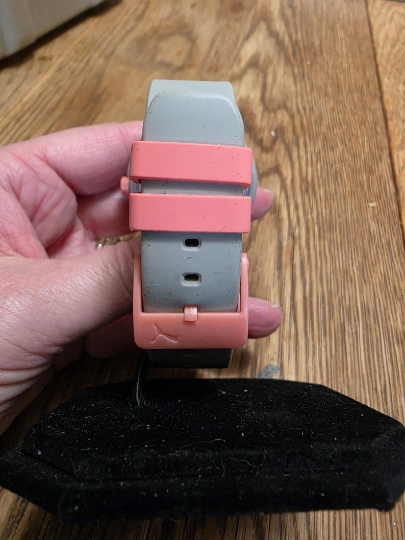 May include: A gray watch band with a pink clasp and straps. The watch band is held over a black velvet display pillow. The watch band is made of a flexible material.