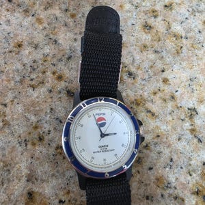 May include: A black Pepsi watch with a white face, blue bezel, and black nylon strap. The watch face displays the Pepsi logo and the text "QUARTZ 3 ATM WATER RESISTANT". The watch has a black case and a black nylon strap.