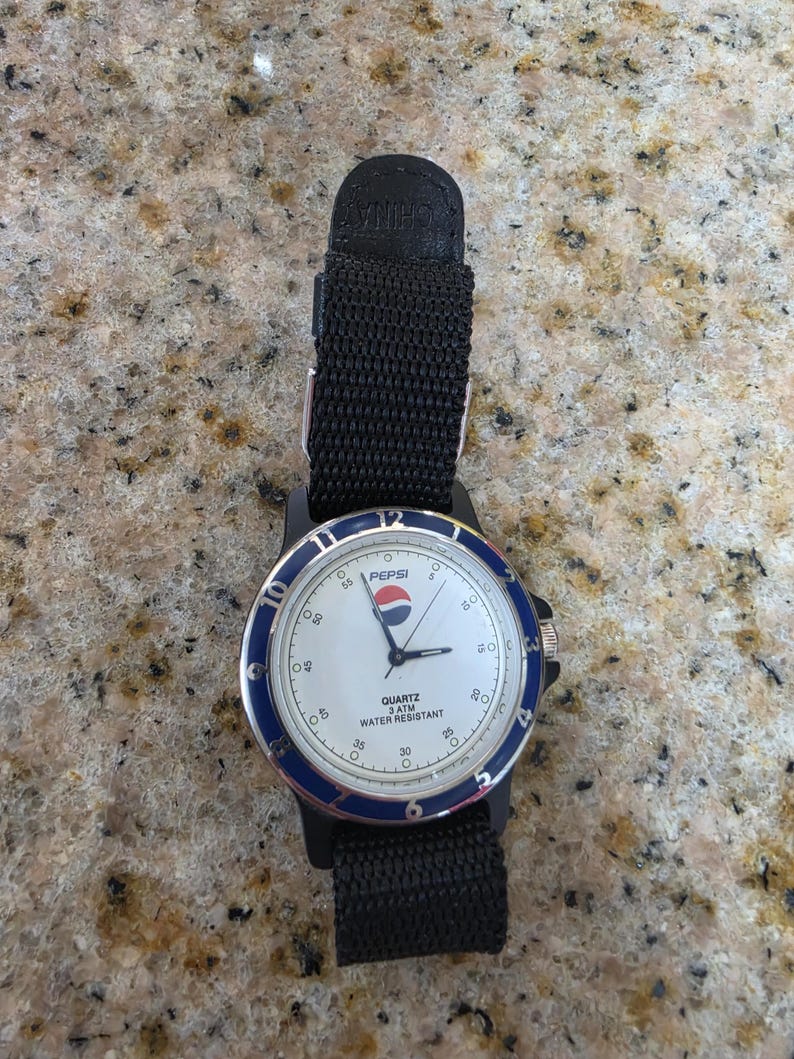 May include: A black Pepsi watch with a white face, blue bezel, and black nylon strap. The watch face displays the Pepsi logo and the text "QUARTZ 3 ATM WATER RESISTANT". The watch has a black case and a black nylon strap.