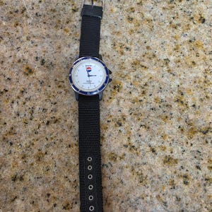 May include: A black wristwatch with a white face, blue bezel, and black nylon strap. The watch face features the Pepsi logo and the words "Quartz Water Resistant". The strap has multiple adjustment holes.