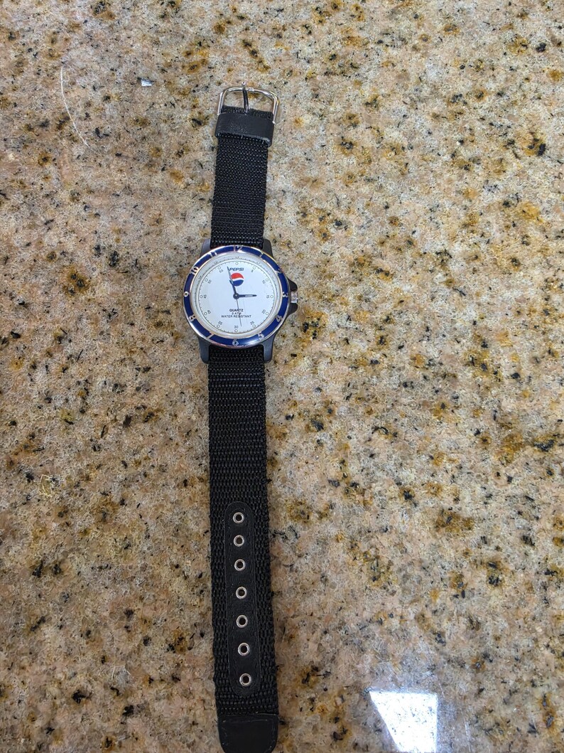 May include: A black wristwatch with a white face, blue bezel, and black nylon strap. The watch face features the Pepsi logo and the words "Quartz Water Resistant". The strap has multiple adjustment holes.