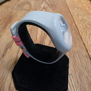 May include: A gray wristwatch with a pink accent on a black display stand. The watch has a clear face with visible internal components. The band is gray with a pink clasp. The watch is displayed on a black, hexagonal stand.