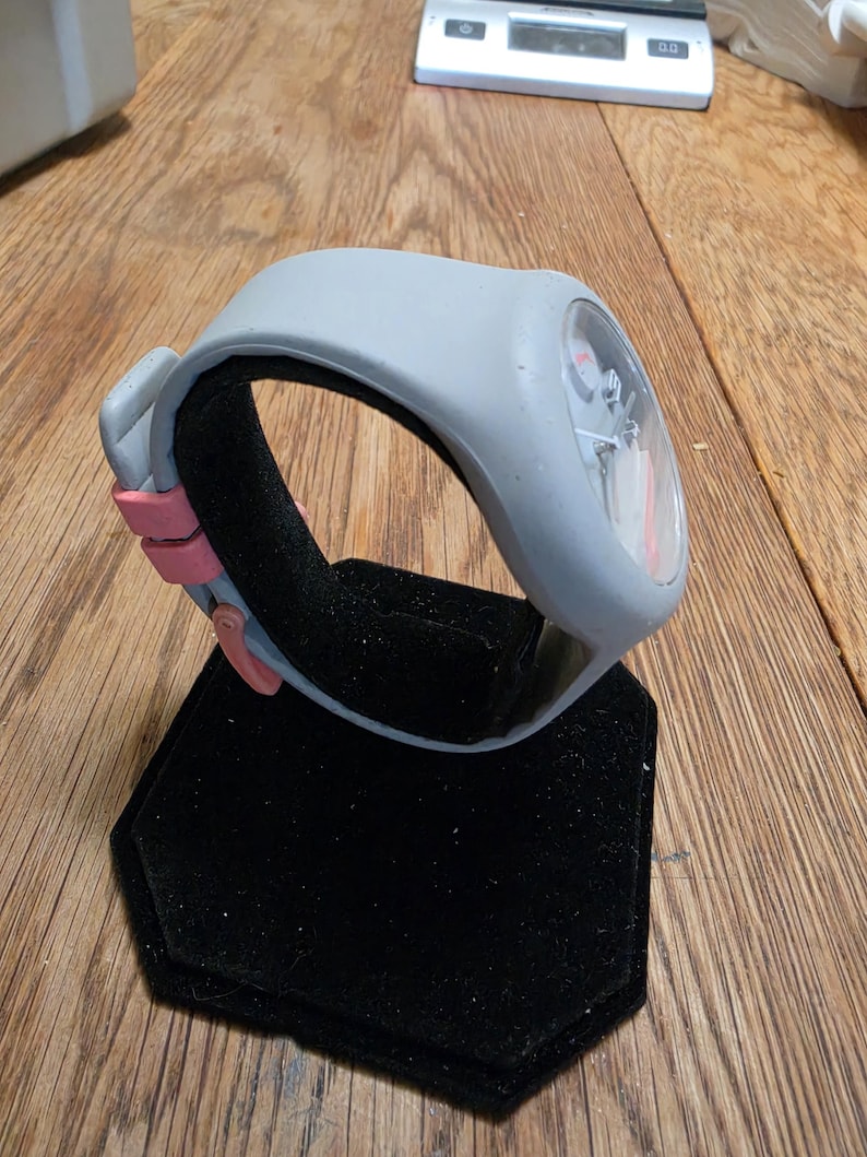 May include: A gray wristwatch with a pink accent on a black display stand. The watch has a clear face with visible internal components. The band is gray with a pink clasp. The watch is displayed on a black, hexagonal stand.