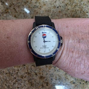 May include: A Pepsi wristwatch with a white face, blue bezel, and black strap. The watch face displays the Pepsi logo and the words "QUARTZ 3ATM WATER RESISTANT". The watch is on a person's wrist.