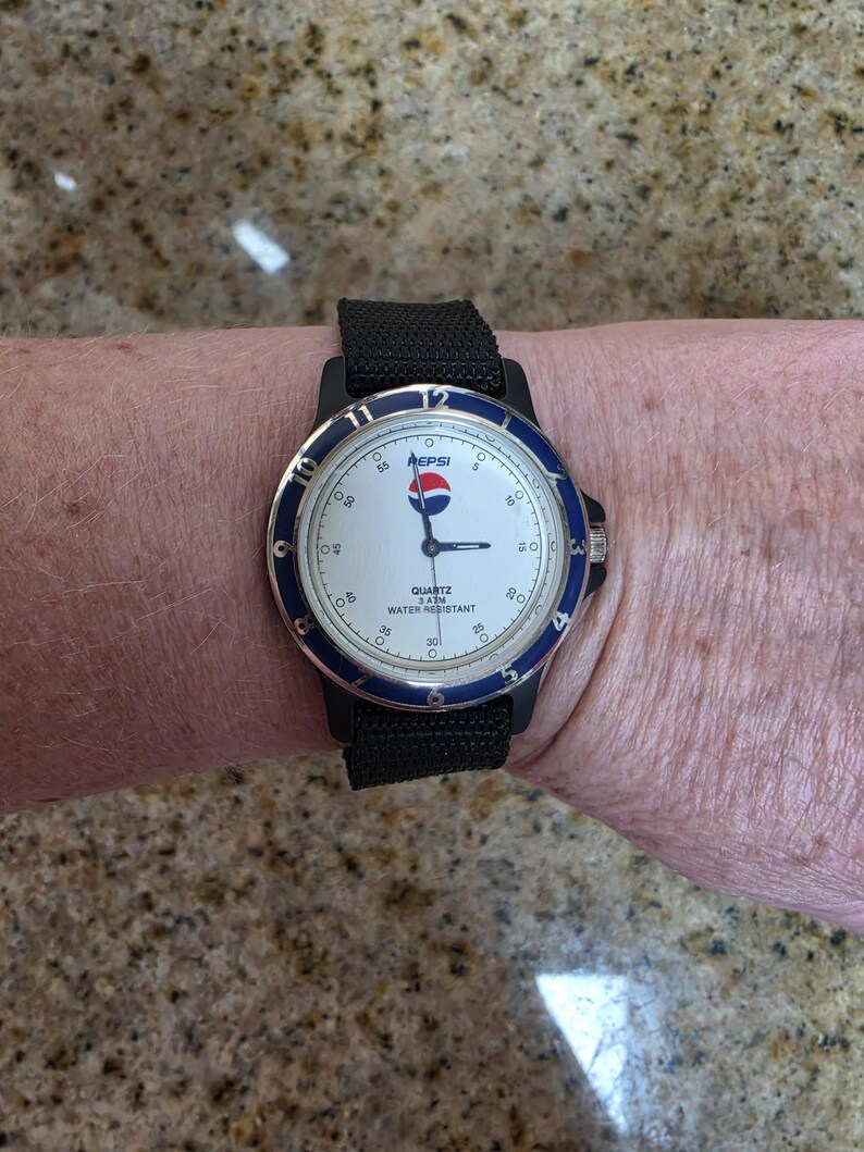 May include: A Pepsi wristwatch with a white face, blue bezel, and black strap. The watch face displays the Pepsi logo and the words "QUARTZ 3ATM WATER RESISTANT". The watch is on a person's wrist.