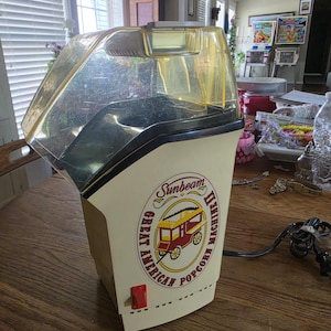 Może przedstawiać: Zabytkowa maszyna do popcornu Sunbeam Great American. Maszyna do popcornu ma kremową podstawę z żółtą i przezroczystą plastikową górą. Maszyna posiada czerwony włącznik/wyłącznik i okrągłe logo z wózkiem do popcornu.