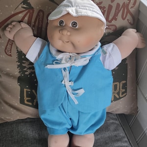 Könnte beinhalten: Eine Vintage-Puppe mit weichem, gebräuntem Gesicht, braunen Augen und einer weißen Mütze. Die Puppe trägt ein hellblaues Outfit mit weißer Krawatte und passenden Schuhen. Die Puppe liegt auf einem Kissen mit Bergmotiv.