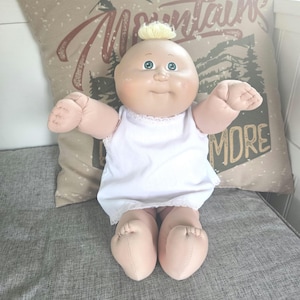 May include: A vintage baby doll with a soft, rounded face, blue eyes, and a small tuft of yellow hair. The doll is wearing a white dress with lace trim. The doll is sitting on a grey surface.