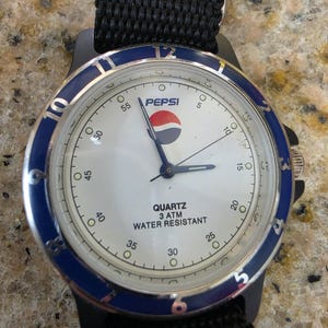 May include: A black wristwatch with a white face and the Pepsi logo. The watch has a blue bezel, a black nylon strap, and the words "QUARTZ 3 ATM WATER RESISTANT" are printed on the watch face.