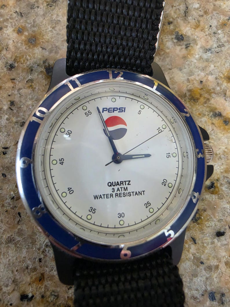 May include: A black wristwatch with a white face and the Pepsi logo. The watch has a blue bezel, a black nylon strap, and the words "QUARTZ 3 ATM WATER RESISTANT" are printed on the watch face.