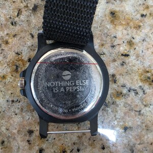 May include: Black watch with a black nylon strap. The watch face features the text "NOTHING ELSE IS A PEPSI" and a Pepsi logo. The watch is water resistant and assembled in China.