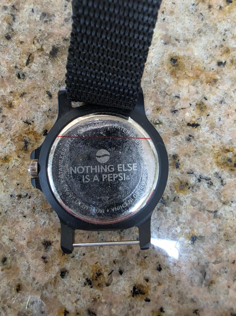 May include: Black watch with a black nylon strap. The watch face features the text "NOTHING ELSE IS A PEPSI" and a Pepsi logo. The watch is water resistant and assembled in China.