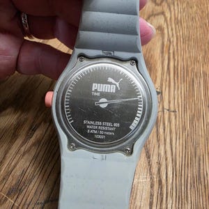 May include: A gray Puma watch with a light gray band. The watch face is silver with the Puma logo and the words "TIME". The back of the watch says "STAINLESS STEEL 805 WATER RESISTANT 5 ATM / 50 meters 103001".