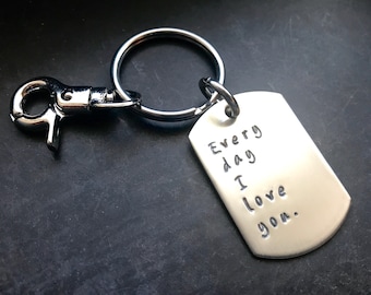 May include: A silver-colored dog tag keychain with the words "Every day I love you." The tag is attached to a metal ring and a clasp. The background is a dark, textured surface.