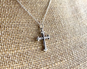 May include: A silver-colored cross pendant necklace. The cross has ornate detailing and hangs from a delicate chain. The necklace is displayed against a textured, neutral-toned background.