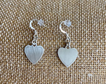 May include: A pair of silver heart-shaped earrings with a brushed finish. The earrings are on a brown textured surface.