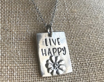 May include: A silver-toned pendant necklace with a rectangular tag. The tag features the words "LIVE HAPPY" in a bold font above a detailed flower design. The necklace chain is also silver-toned, creating a simple yet elegant accessory.