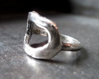 Pode incluir: Um anel de prata com um design aberto único. O anel é feito de metal e tem um acabamento liso.