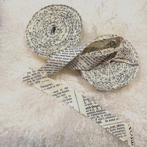 May include: Two rolls of decorative paper ribbon with black text on an off-white background. One roll is partially unraveled, revealing a strip of the same paper. The ribbon is tied with twine, suggesting a craft or scrapbooking theme.