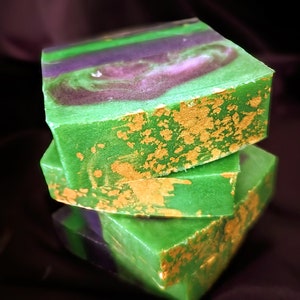 May include: Three stacked, square-shaped soaps. The soaps are primarily green with purple and white swirls. Gold flecks adorn the edges. The soaps are arranged against a dark, blurred background. The soaps appear to be handmade.